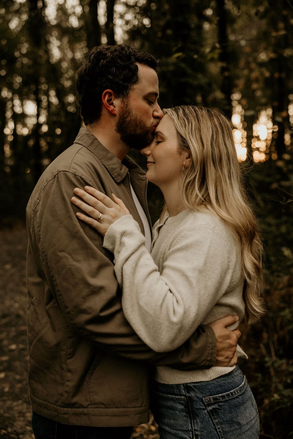 A romantic couple shares a tender embrace in a moody forest setting during golden hour.