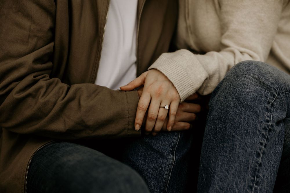 Close-up view of hands resting on denim jeans, wearing brown coat and cream-colored sweater.