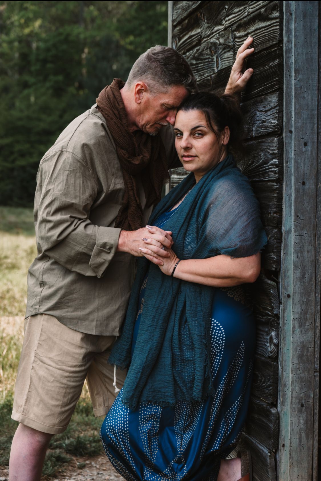A middle-aged couple shares an intimate moment leaning against a rustic stone wall outdoors, surrounded by lush green trees.