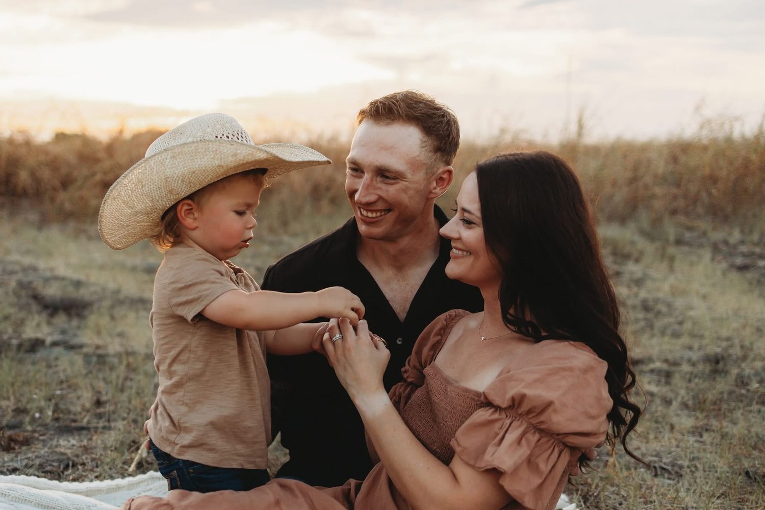 Young couple with toddler boy in cowboy hat sharing tender moment in golden field at sunset.