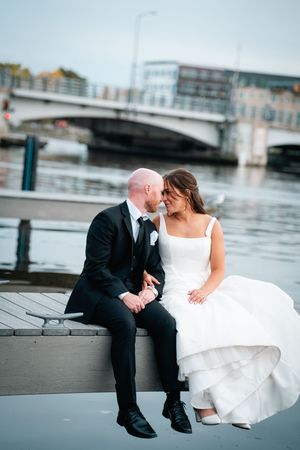 A romantic wedding couple sits together on a dock by the water with a bridge in the background at dusk.