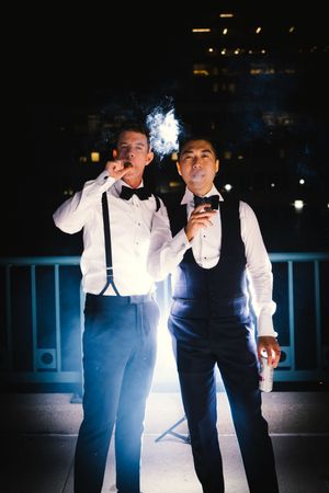 Two people in formal attire with suspenders and bow ties smoke together at night on a balcony with city lights behind them.