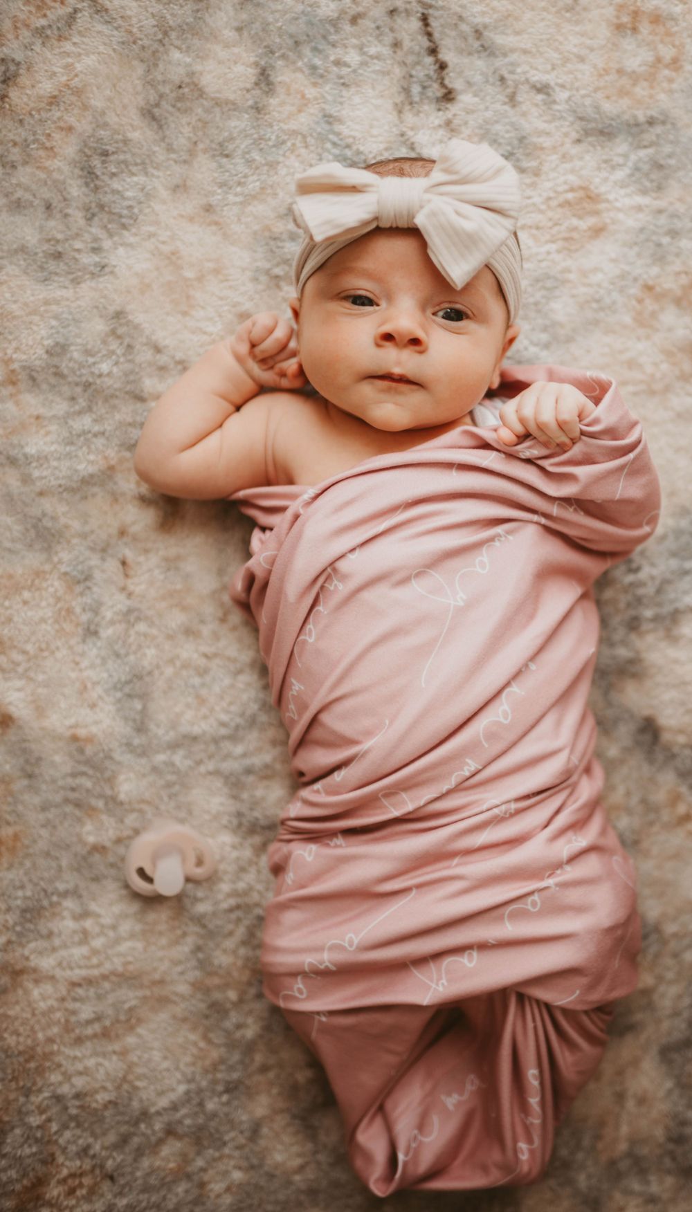 Newborn baby wearing pink outfit and white bow lying on textured backdrop.