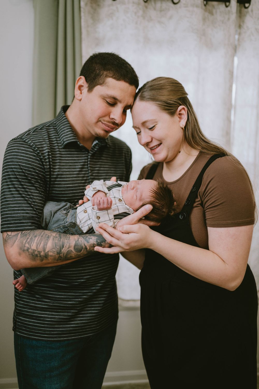 New parents smiling and cradling their newborn baby together indoors, father with tattoos wearing striped shirt.