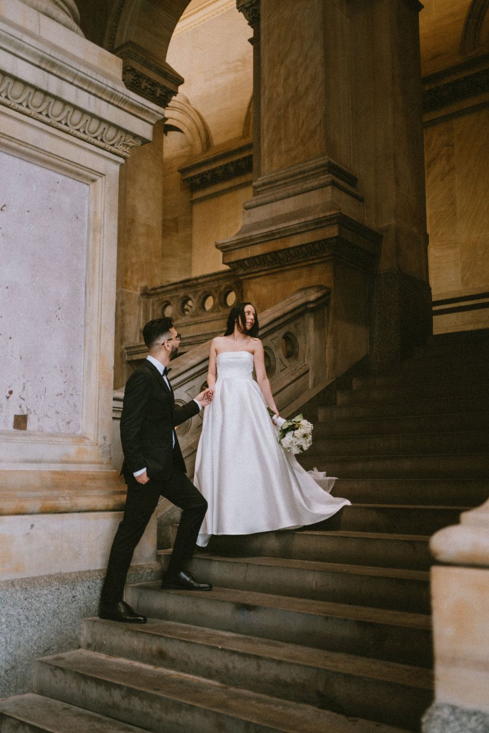 Bride and groom on grand stone staircase, bride in strapless white gown holding bouquet, groom in black suit.
