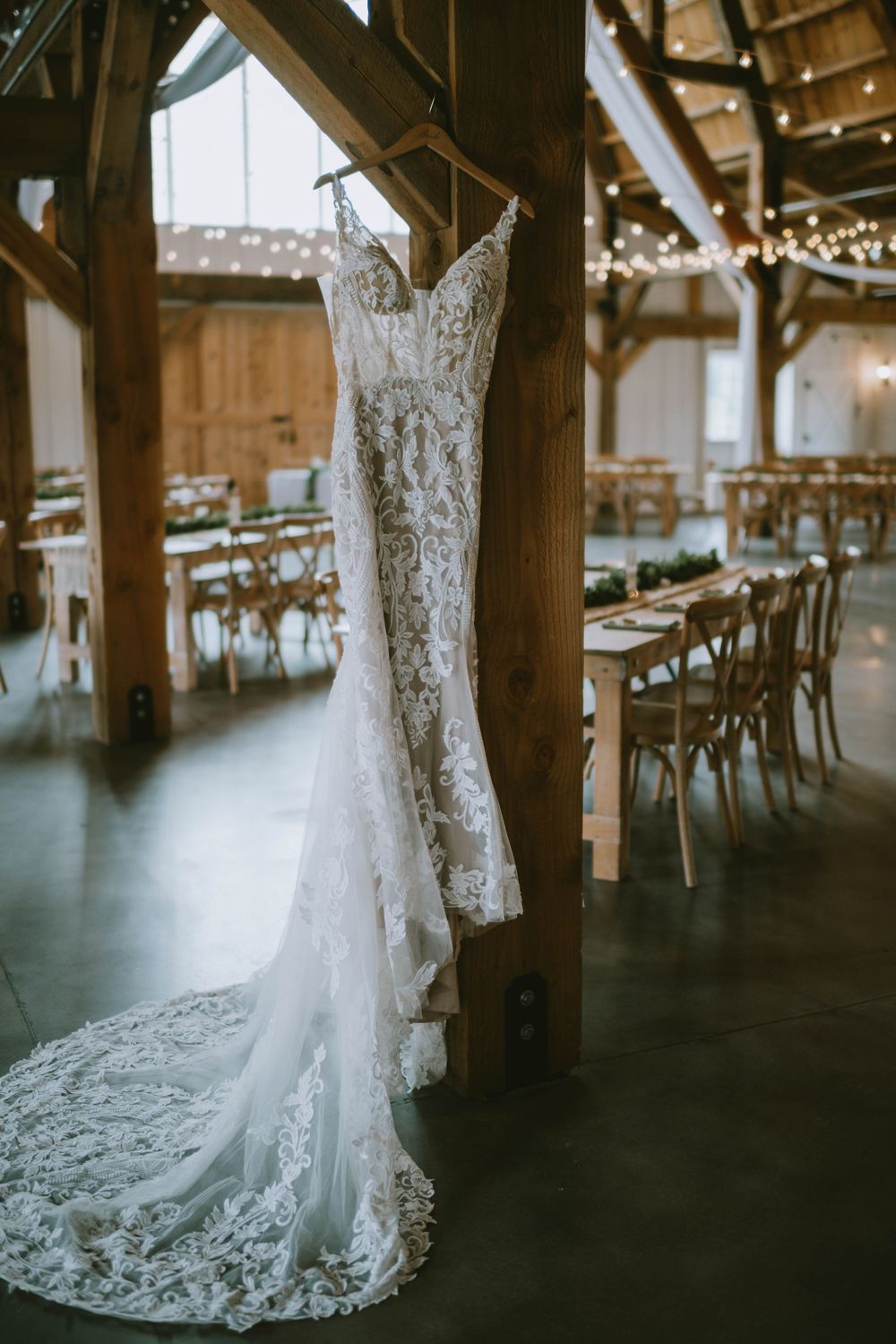 Elegant lace wedding dress hanging in a rustic barn venue with string lights and reception tables in the background.