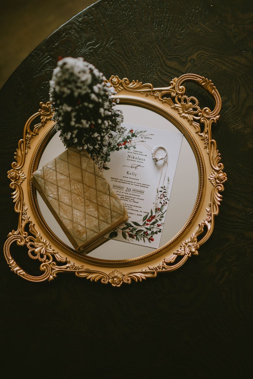 Gold ornate mirror tray with wedding invitation, diamond ring, floral bouquet, and vintage book on dark table.