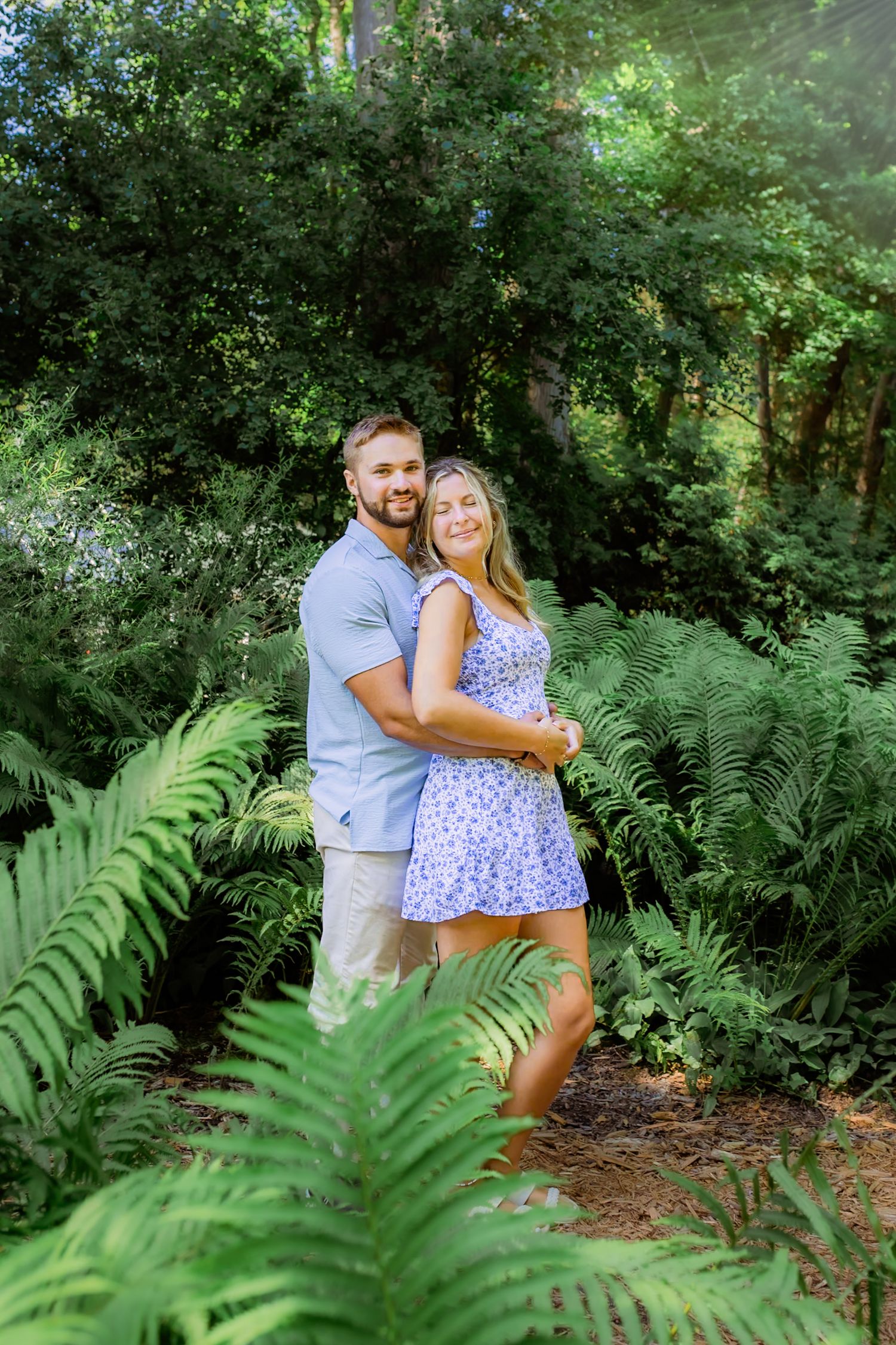 A couple embraces and smiles among lush green ferns and trees in an outdoor garden setting.