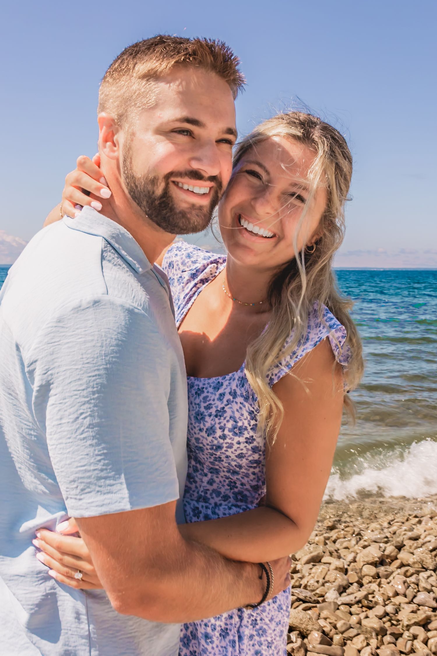 Happy couple embracing and smiling on a rocky beach with turquoise ocean waves in the background on a sunny day.