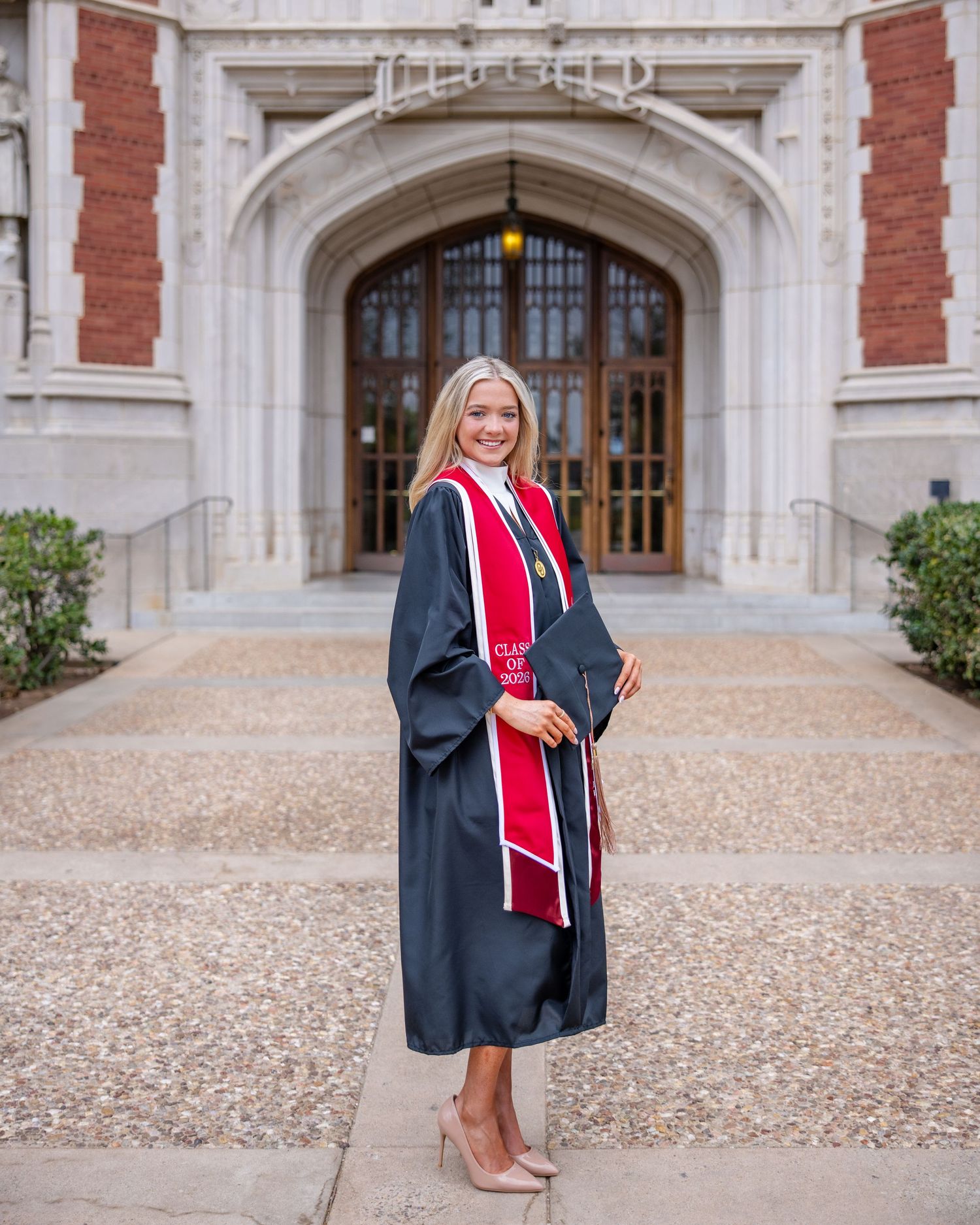Graduate in cap and gown with graduation stole stands in front of ornate university building entrance archway.