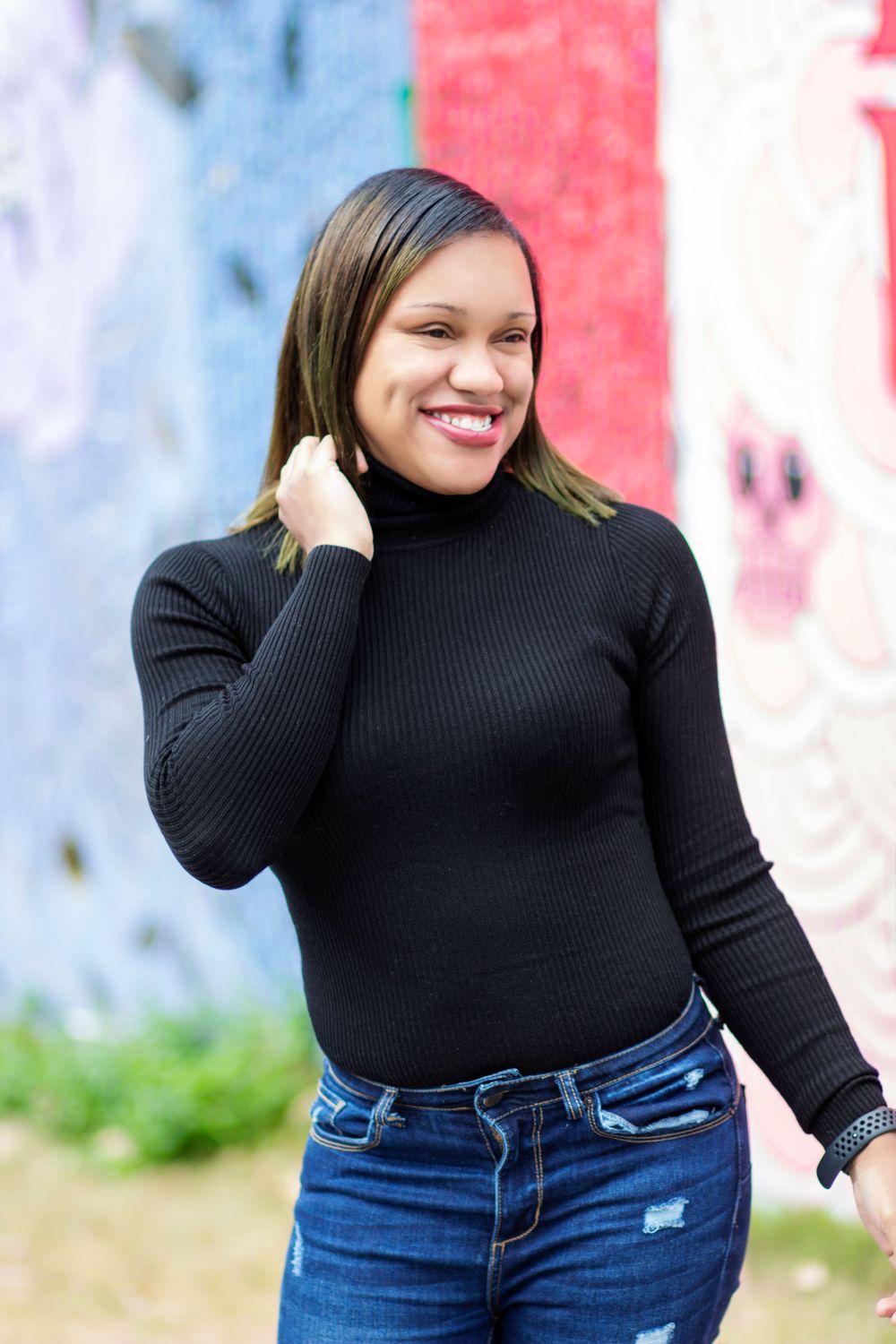 Plus size model poses in black turtleneck and jeans against colorful wall background.