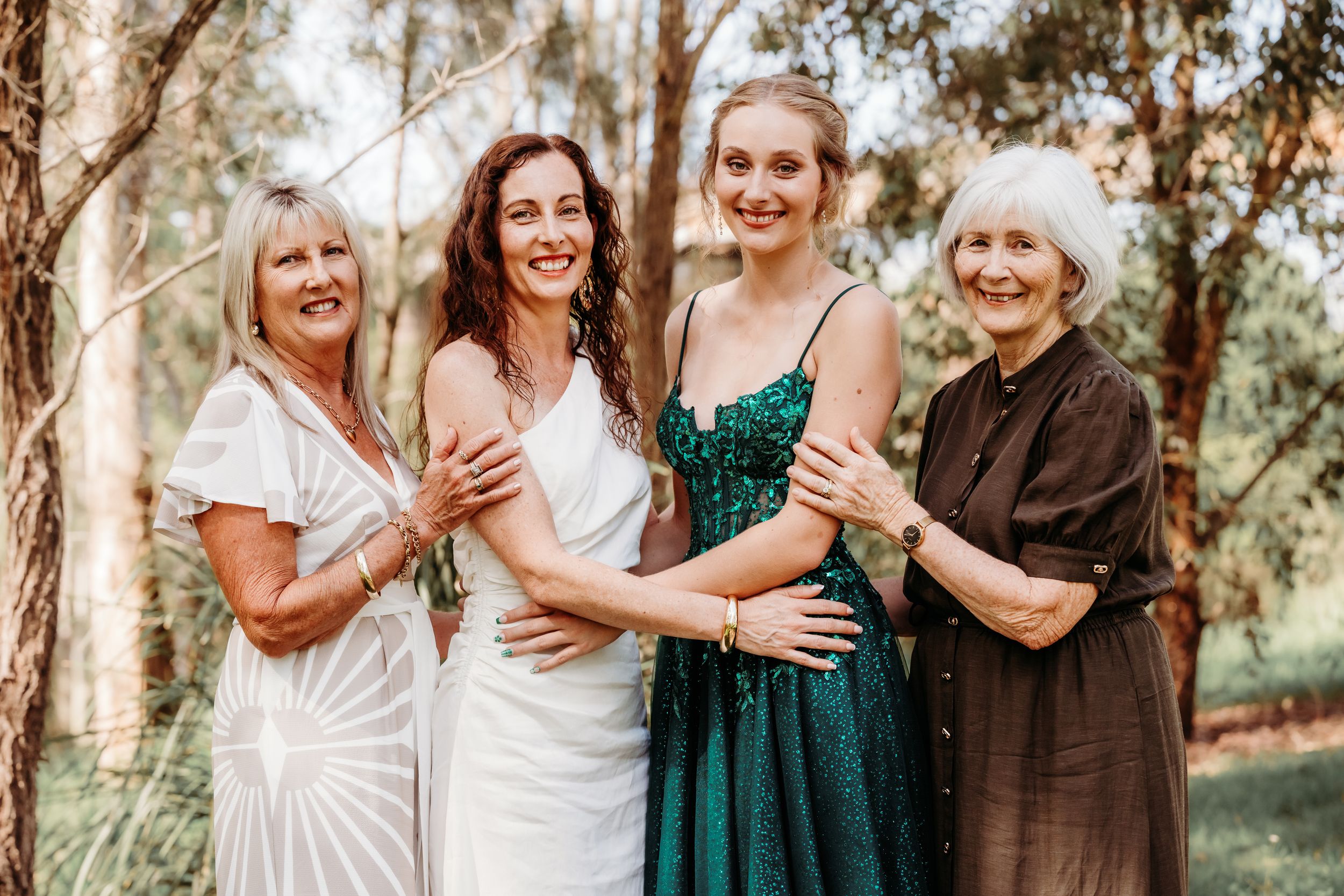 A group of women in white and emerald green dresses stand together laughing outdoors in a natural setting.