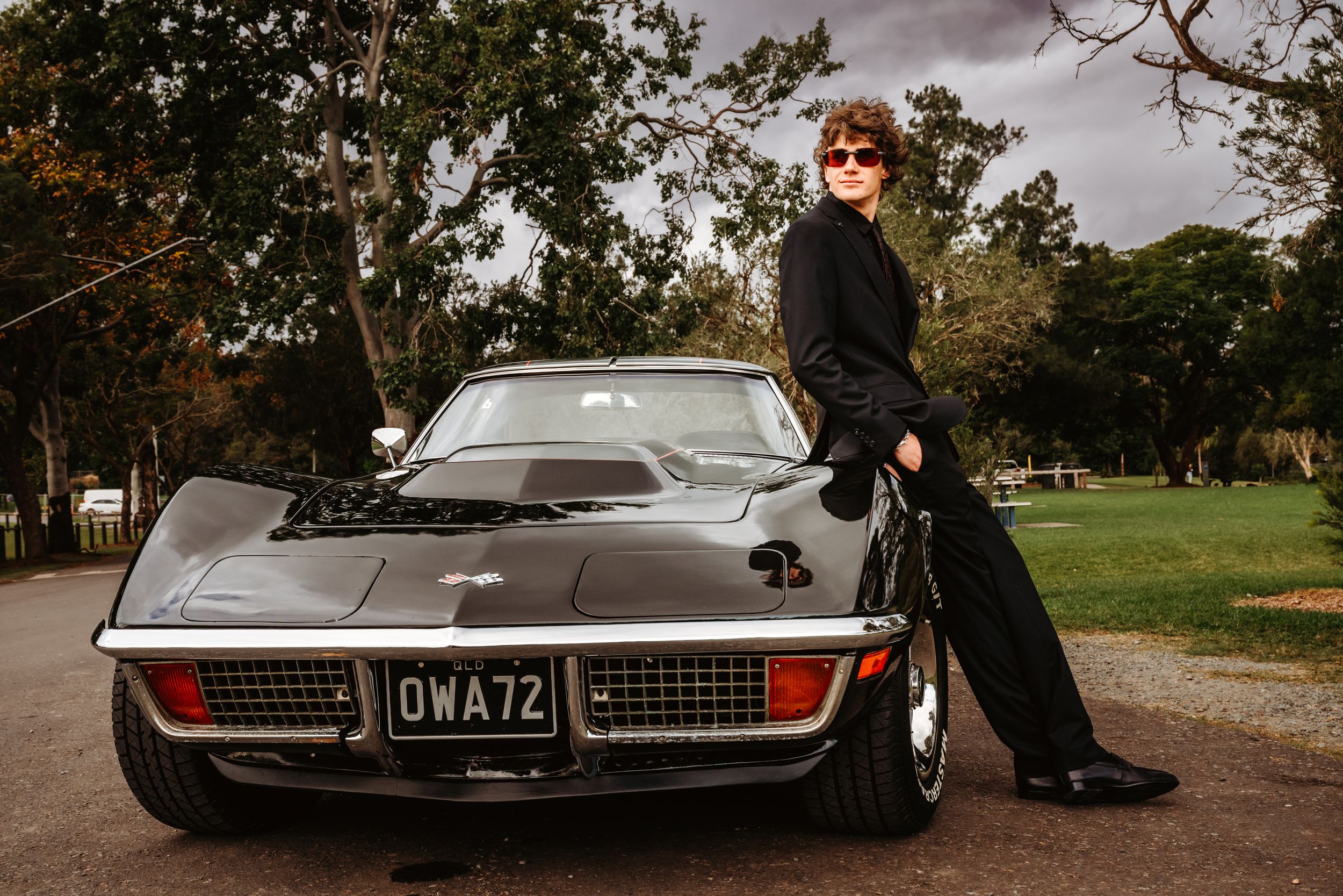 Person in black outfit leaning against a classic black Corvette sports car with license plate OWA72 outdoors.