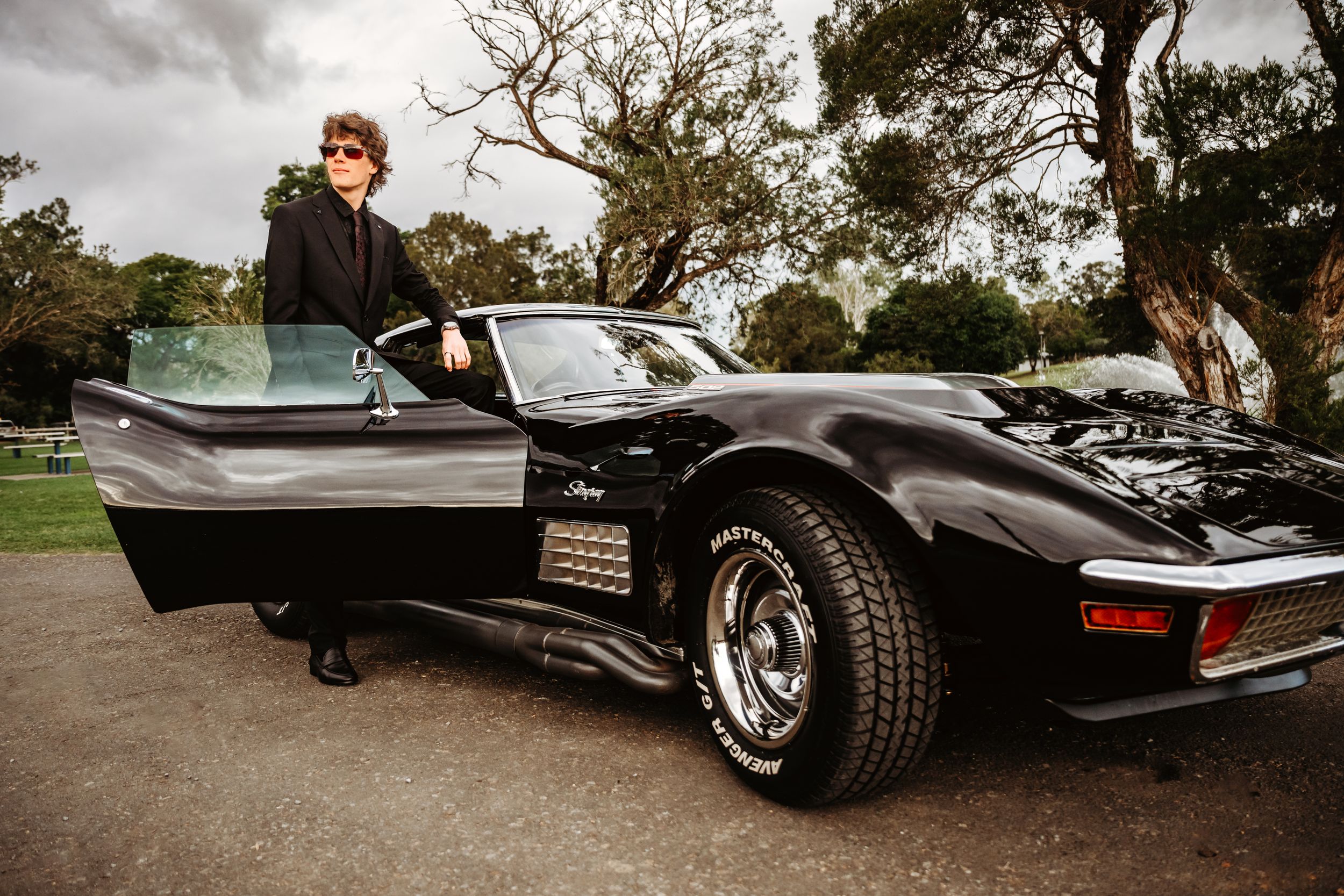 Person in black outfit posing beside a classic black Corvette convertible outdoors.