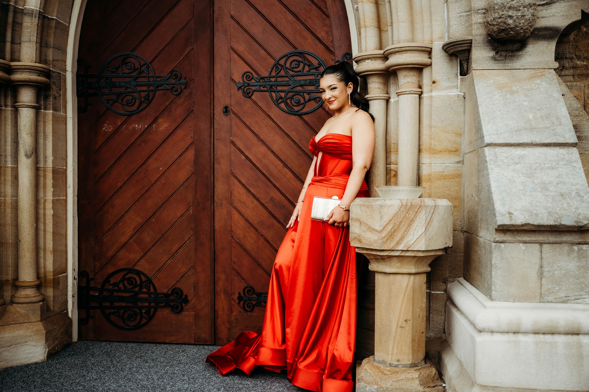 A person in an elegant red formal gown poses against a stone church entrance with ornate wooden doors.