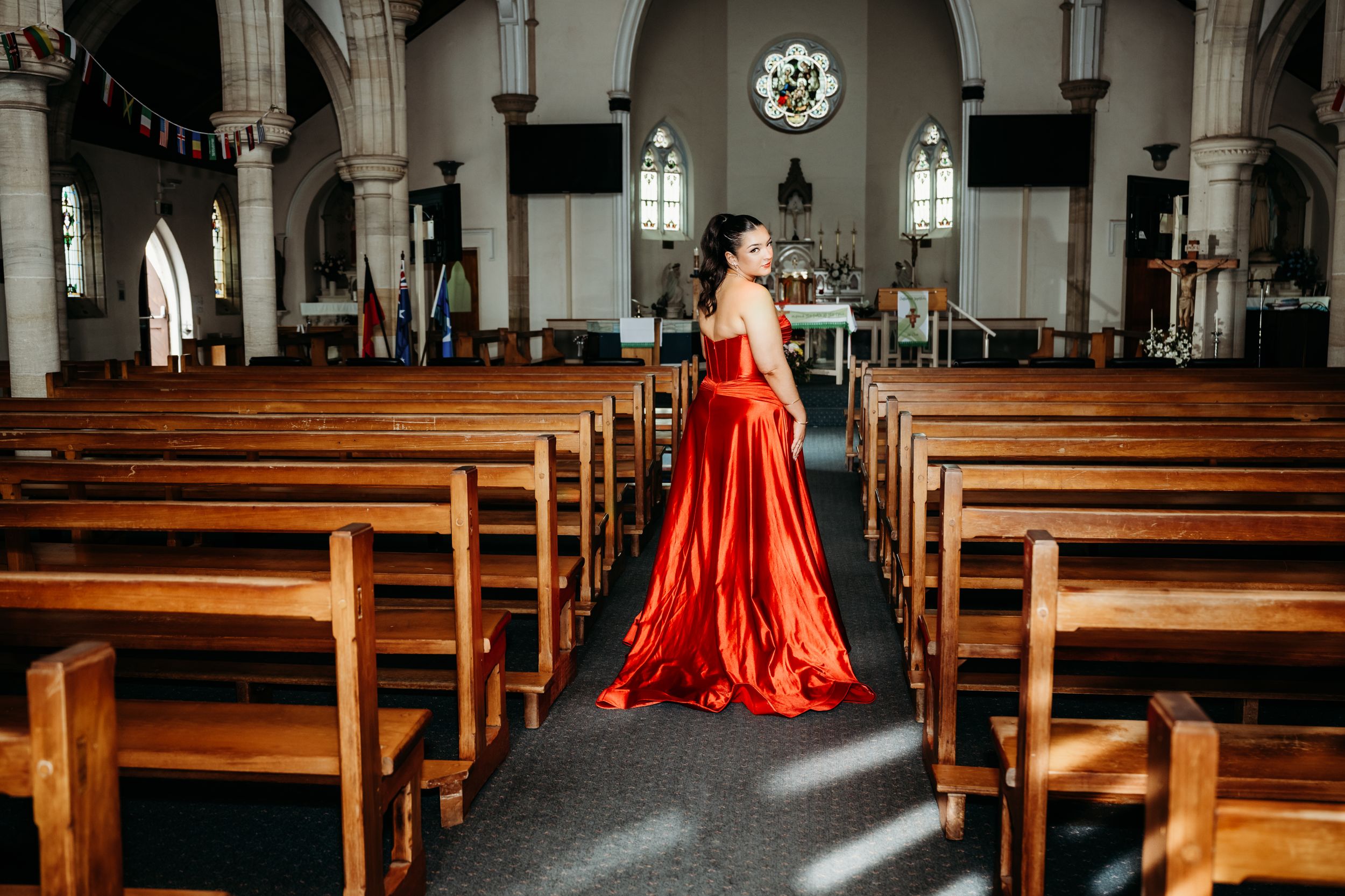 A person in a flowing red evening gown poses elegantly in a church with wooden pews and stained glass windows.