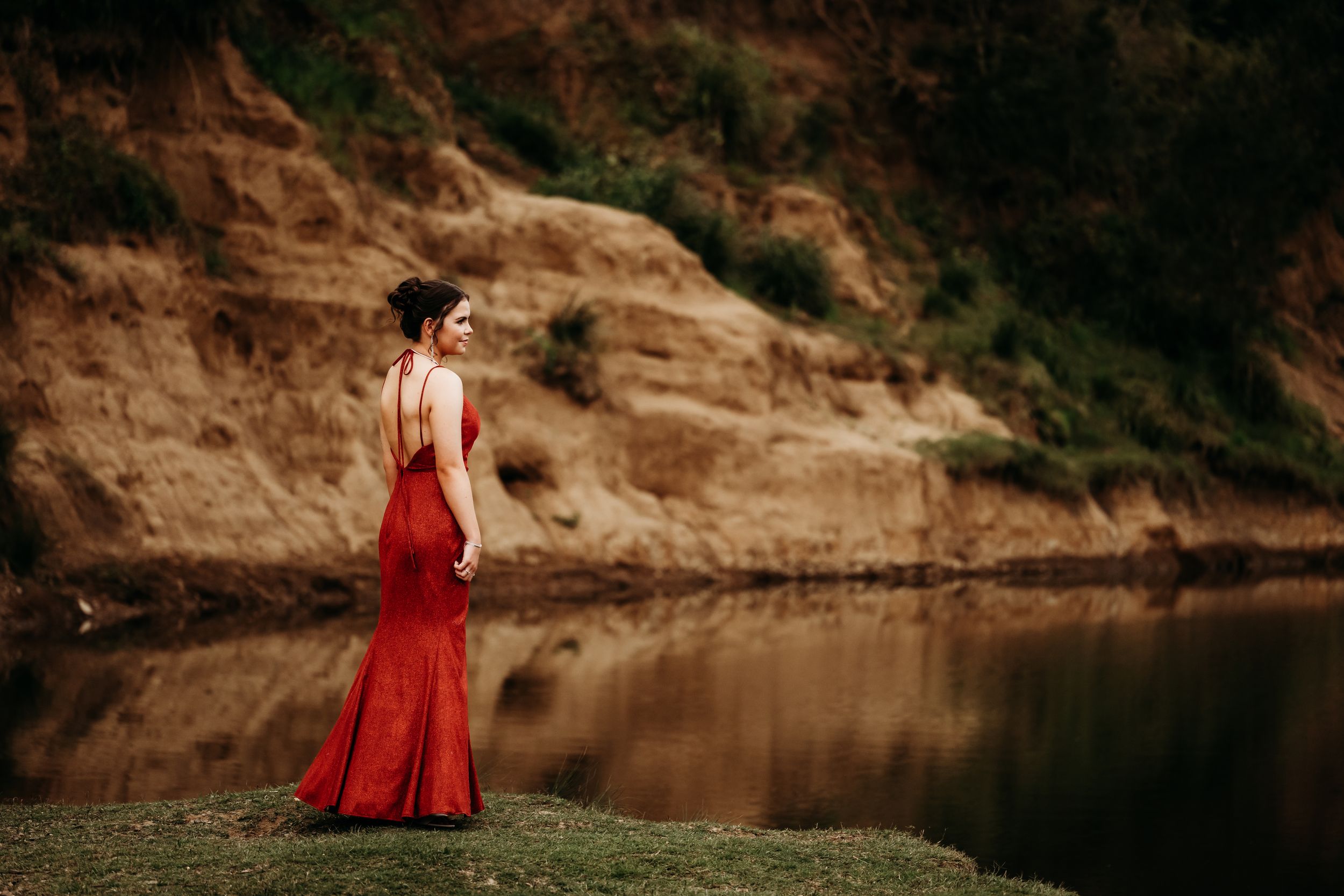 Someone in a flowing red dress stands elegantly by a serene lake with rocky cliffs in the background.