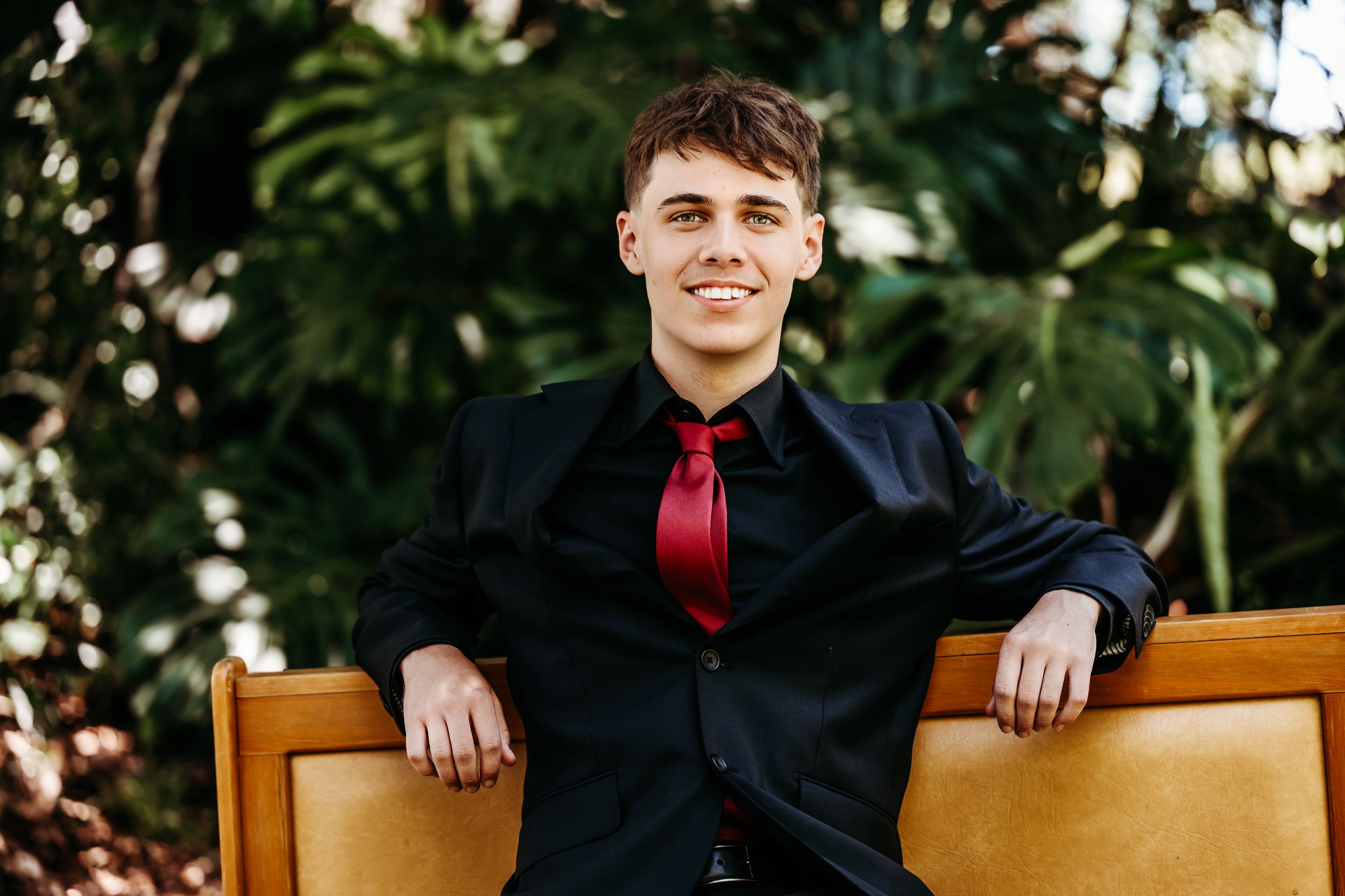 A person in a black dress shirt and red tie sits on a wooden chair with tropical foliage in the background.