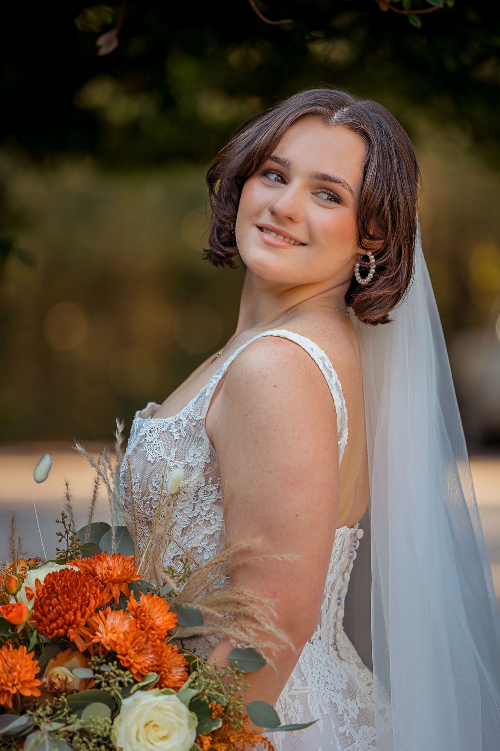 A bride in a white lace gown holds an orange and cream floral bouquet while posing outdoors during golden hour.