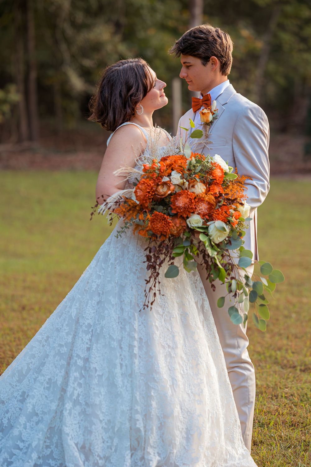 A romantic outdoor wedding photo with a stunning white lace gown and vibrant orange and white floral bouquet.