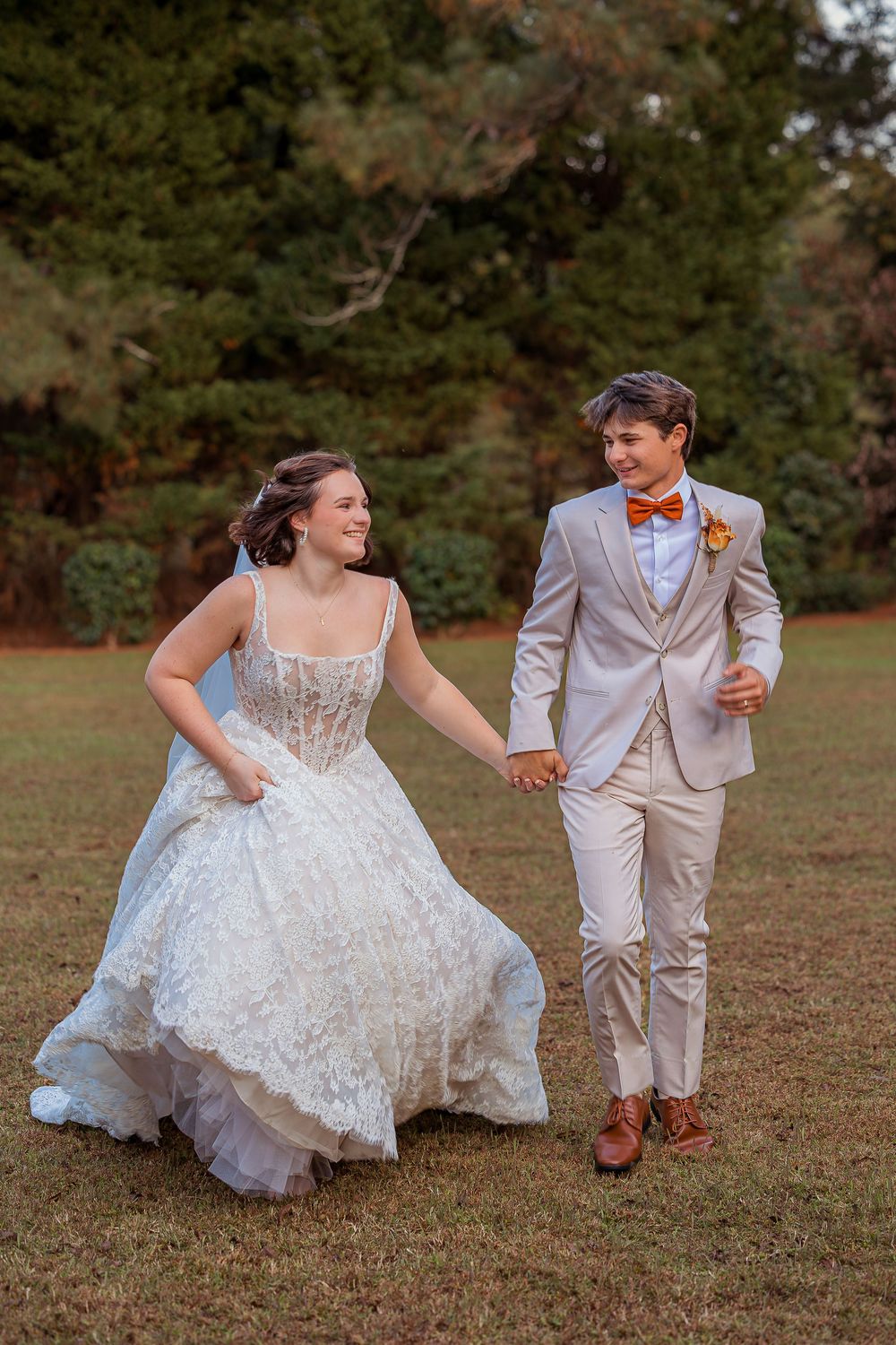A couple in wedding attire playfully poses together in a grassy field during golden hour.