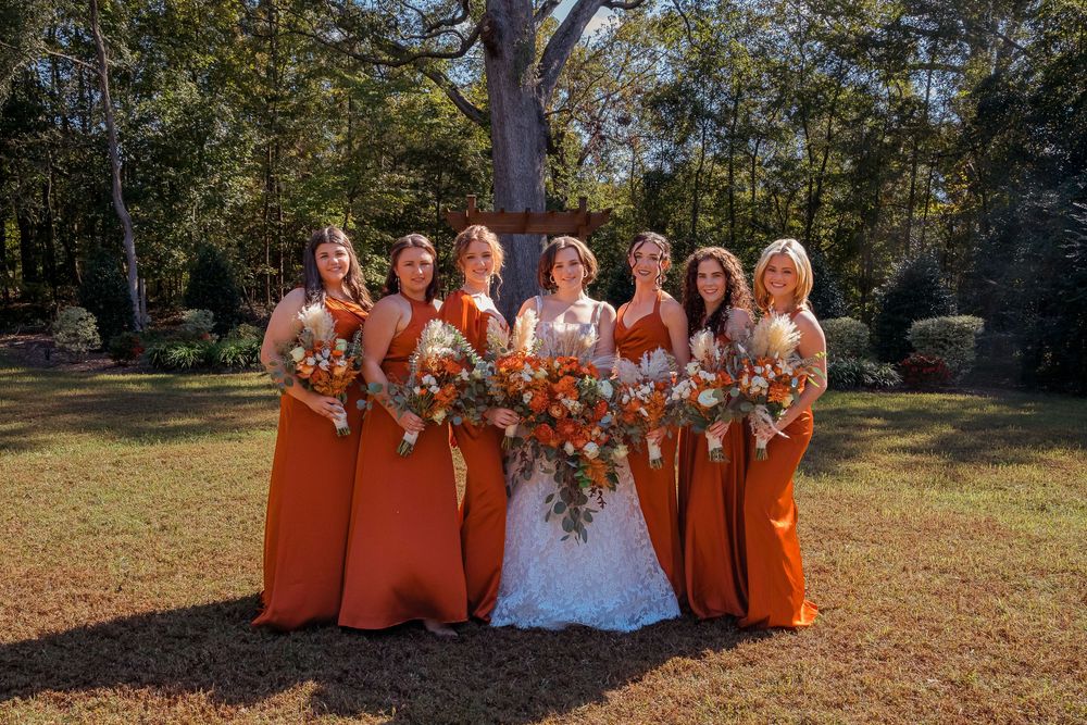 Wedding party wearing bright orange dresses poses outdoors in autumn setting with matching floral bouquets.