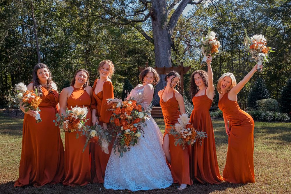 A bridal party poses outdoors with bridesmaids in burnt orange dresses alongside a bride in white.