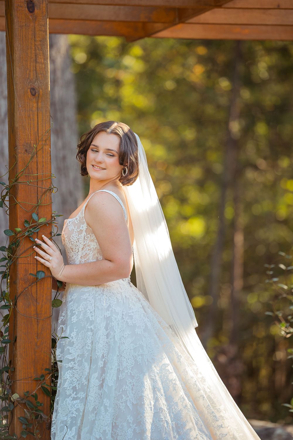 A bride in a white lace wedding dress poses gracefully in a rustic wooden gazebo during golden hour.
