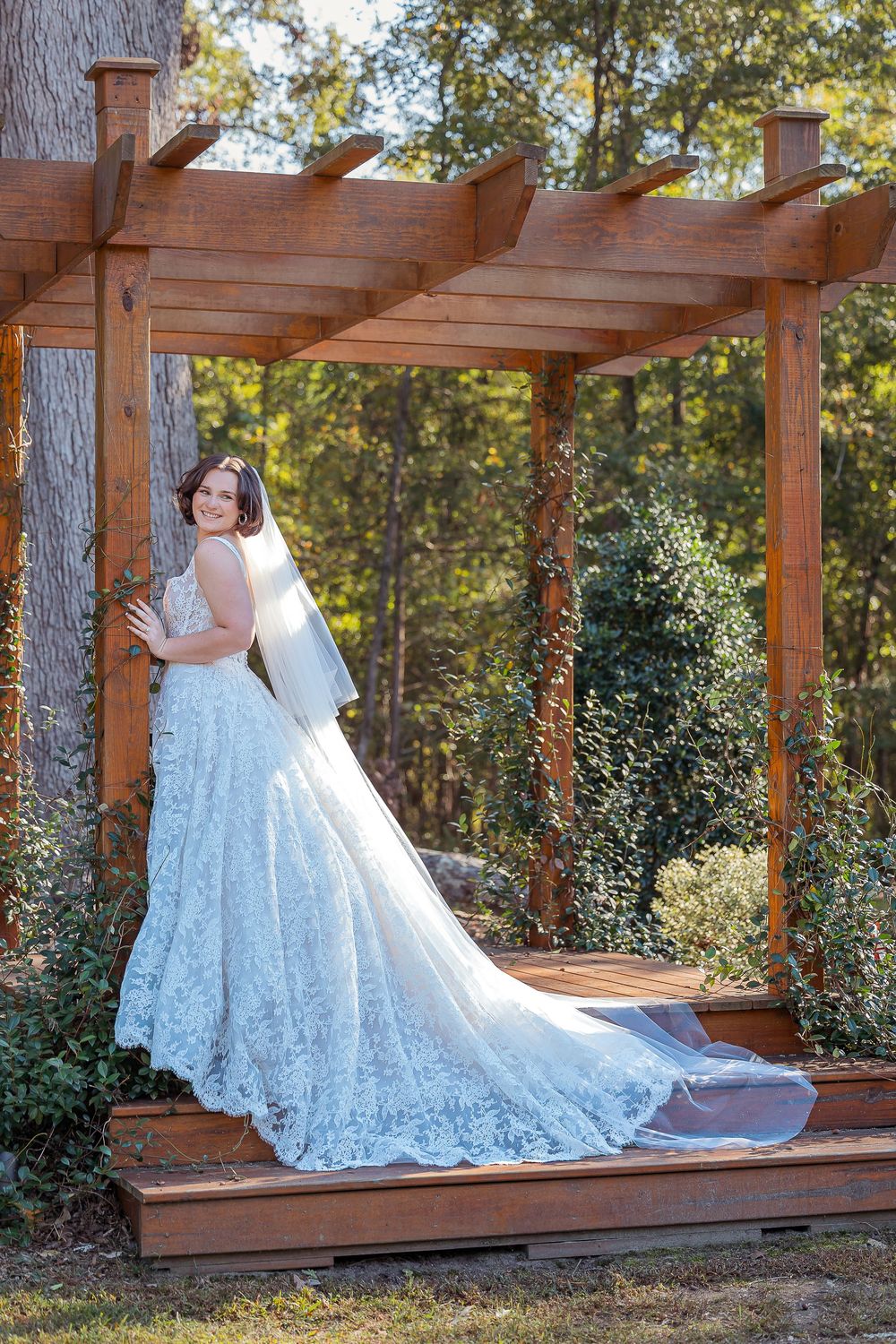 Stunning white lace wedding dress with long train photographed in outdoor wooden pergola setting during golden hour.