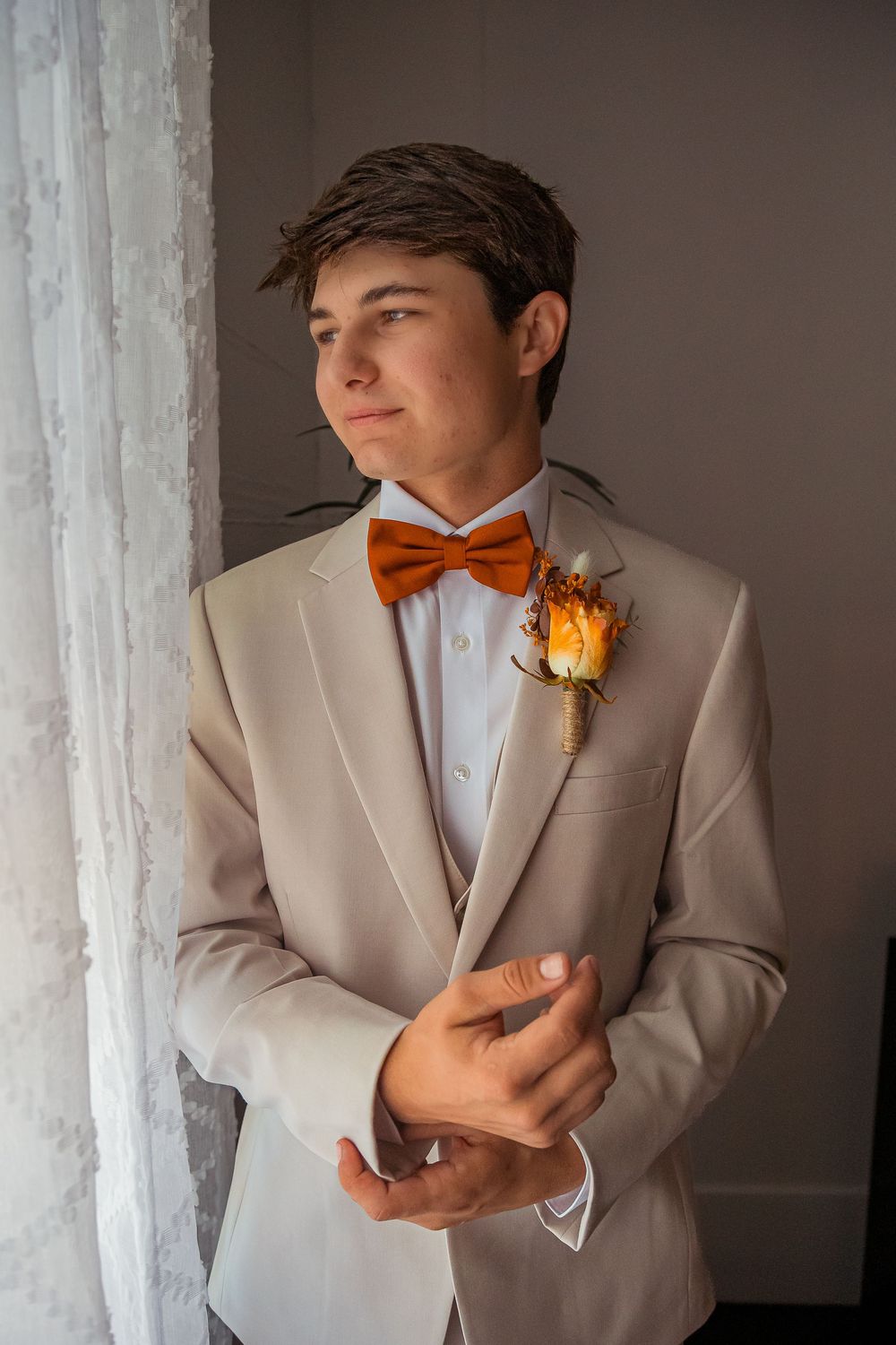 A groom in a beige suit and orange bowtie poses against a light colored wall in an artistic portrait series.
