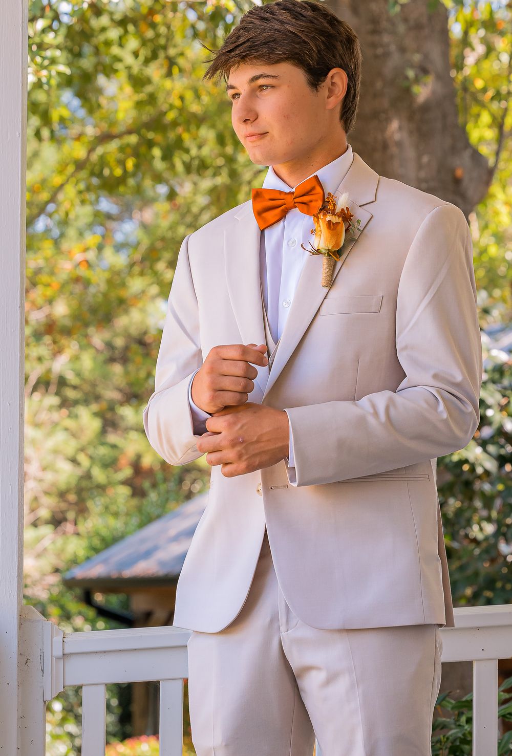 A person in a light gray suit with orange bow tie and boutonniere poses outdoors in natural lighting.