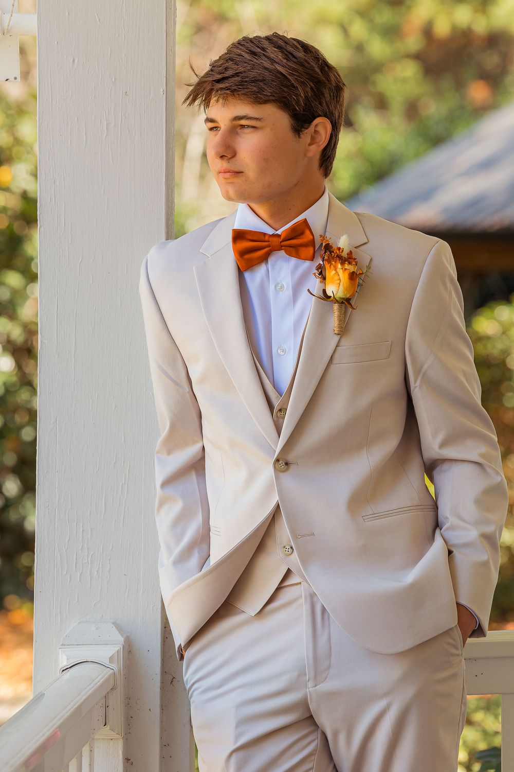 A formal wedding attire featuring a tan suit with orange bow tie and boutonniere against an outdoor background.