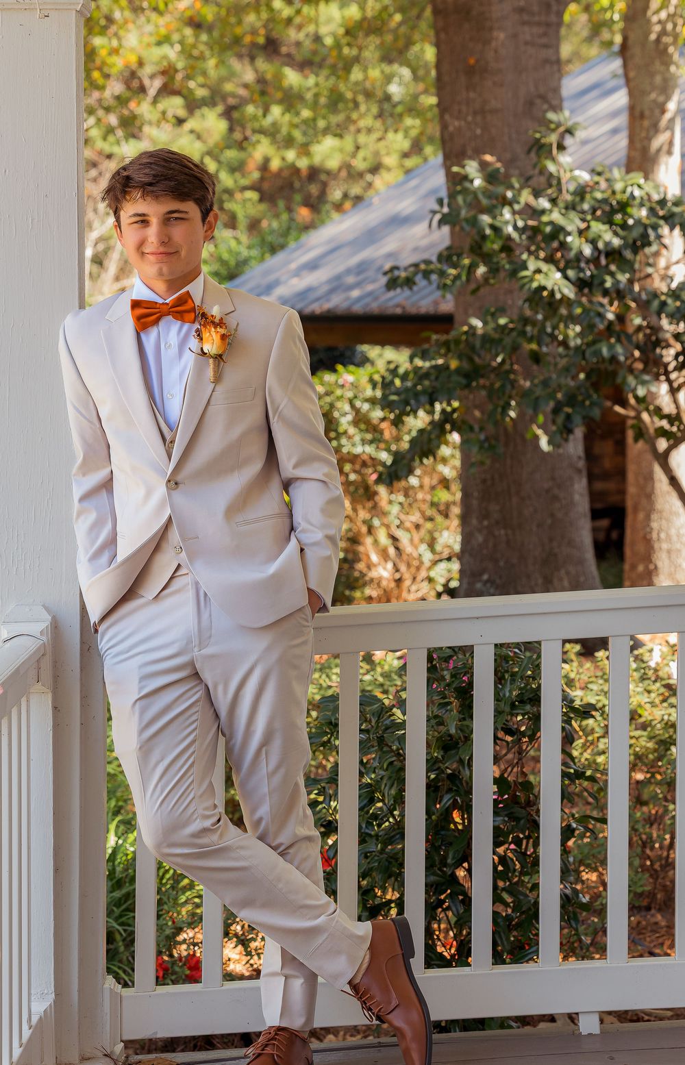 A groom in tan suit and orange bowtie poses on a white porch railing with autumn foliage in background.