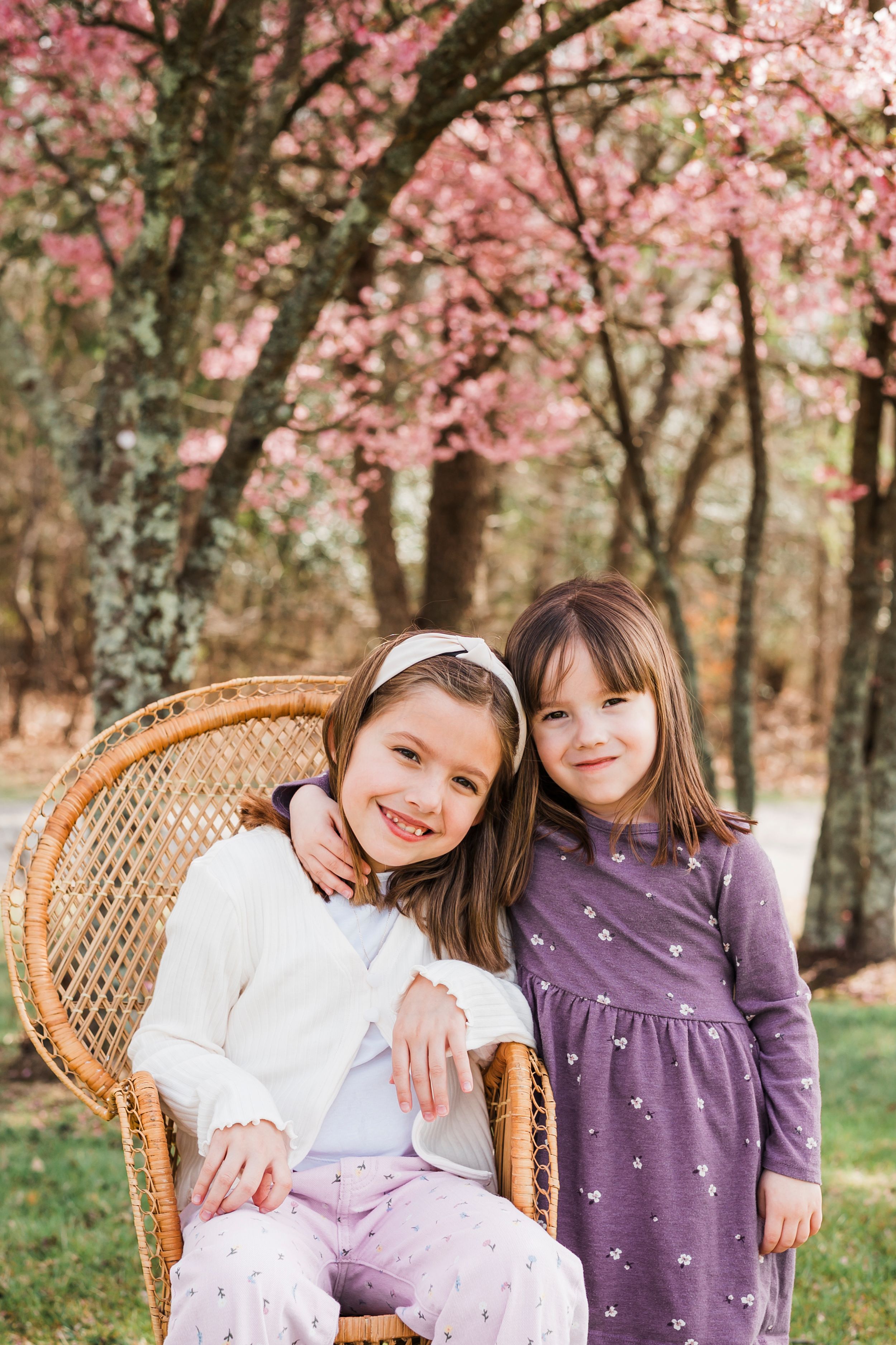 Two children pose together near cherry blossom trees, one sitting in a woven peacock chair wearing white and pink.