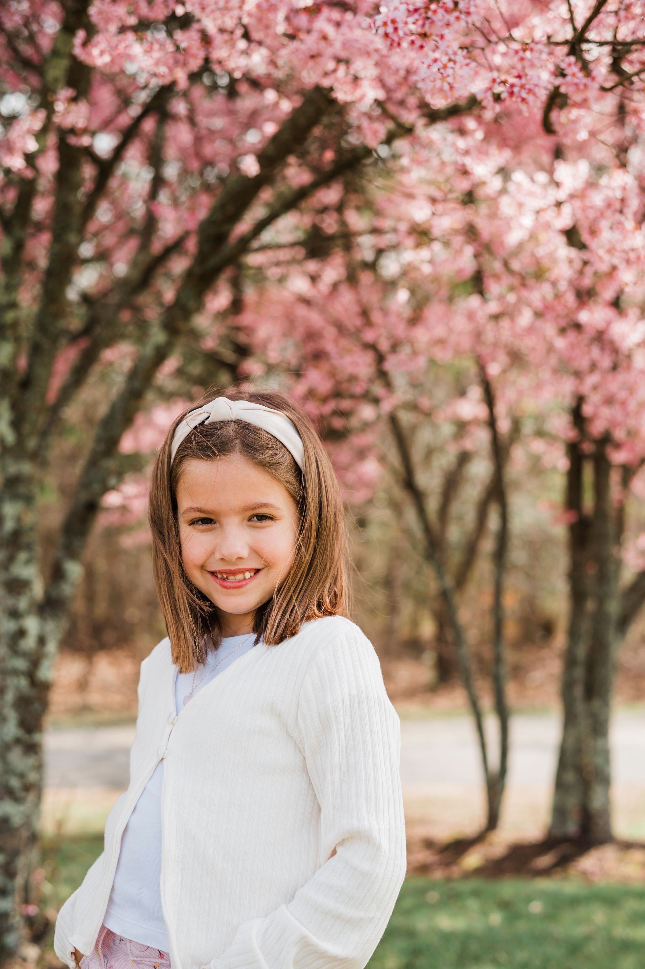 A springtime portrait under blooming pink cherry blossoms with a white sweater and headband.