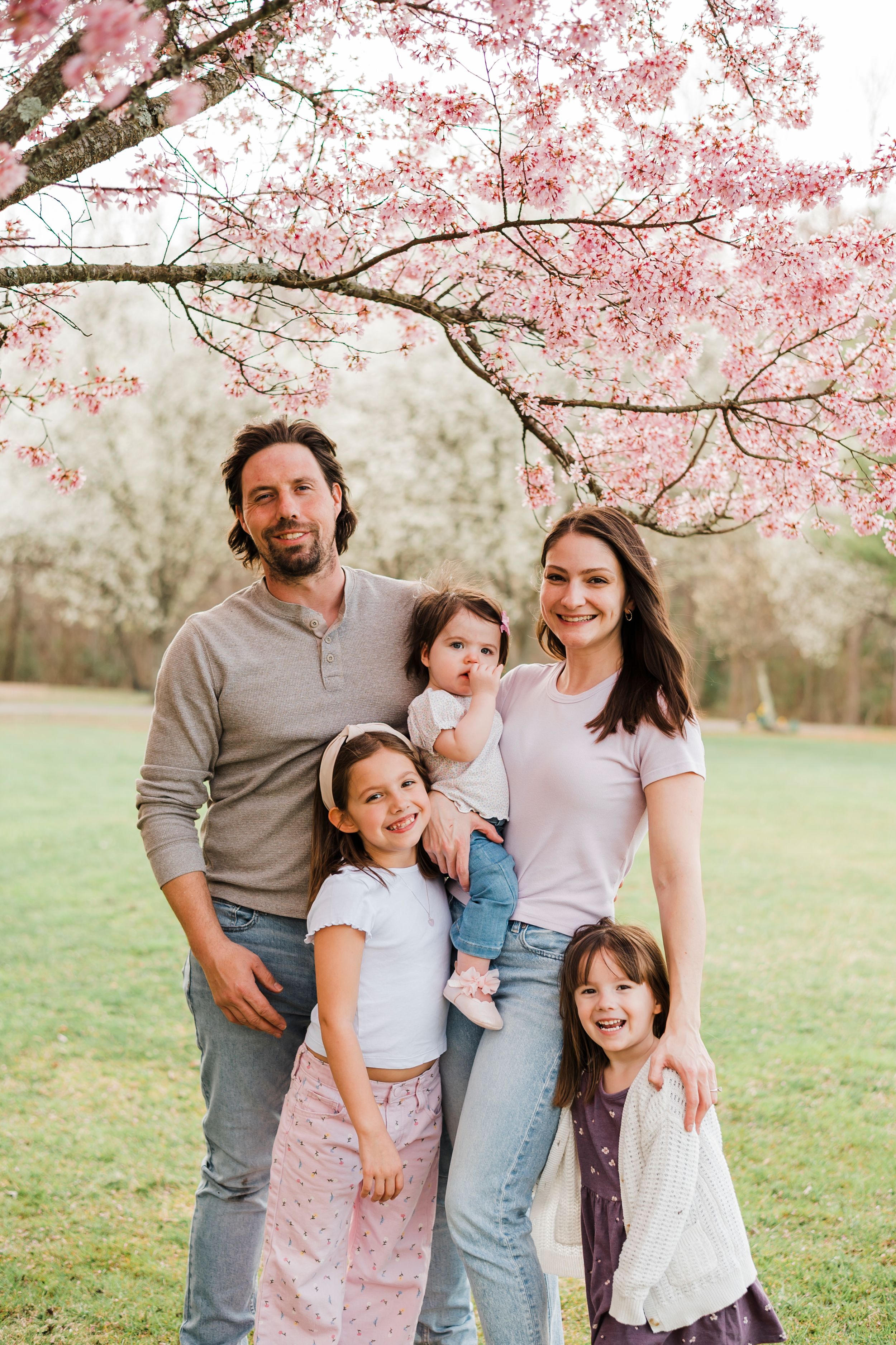 Family standing together under blooming cherry blossoms in grassy field during springtime photo session.