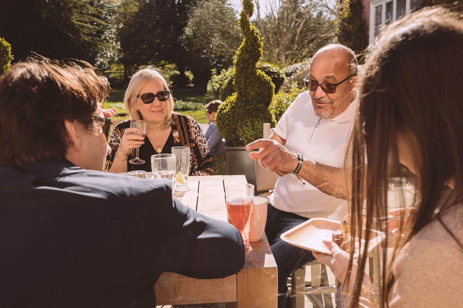Group gathers for outdoor dining and drinks at sunny restaurant patio on warm afternoon.