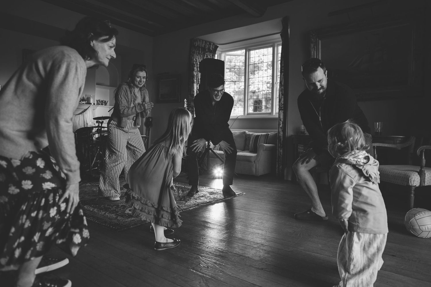 A black and white photograph shows people dancing together in a rustic room with sunlight streaming through a window.