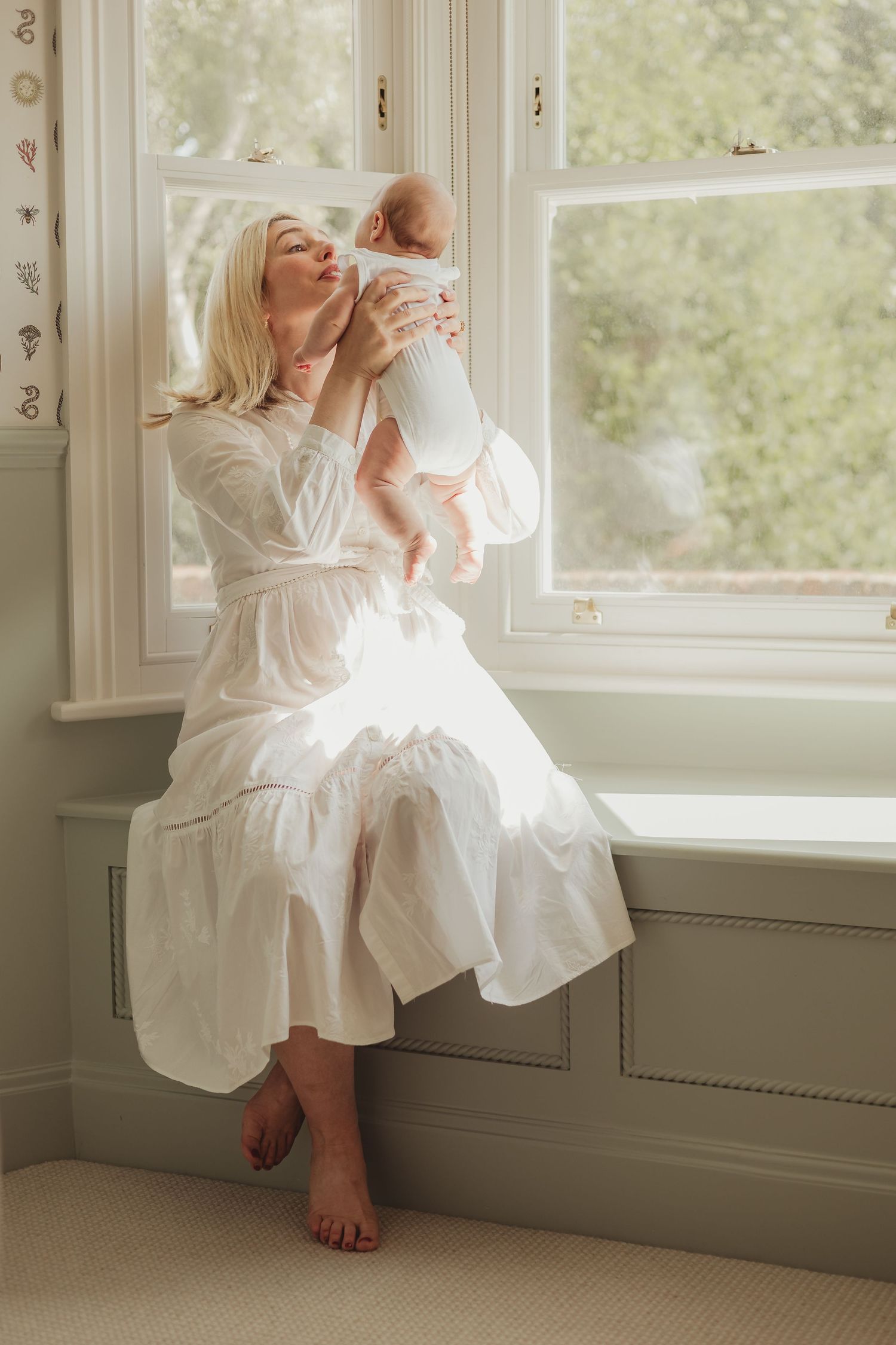 A smiling mother, dressed in white, lifts up her baby to give her a kiss, bathed in natural light from the bay window