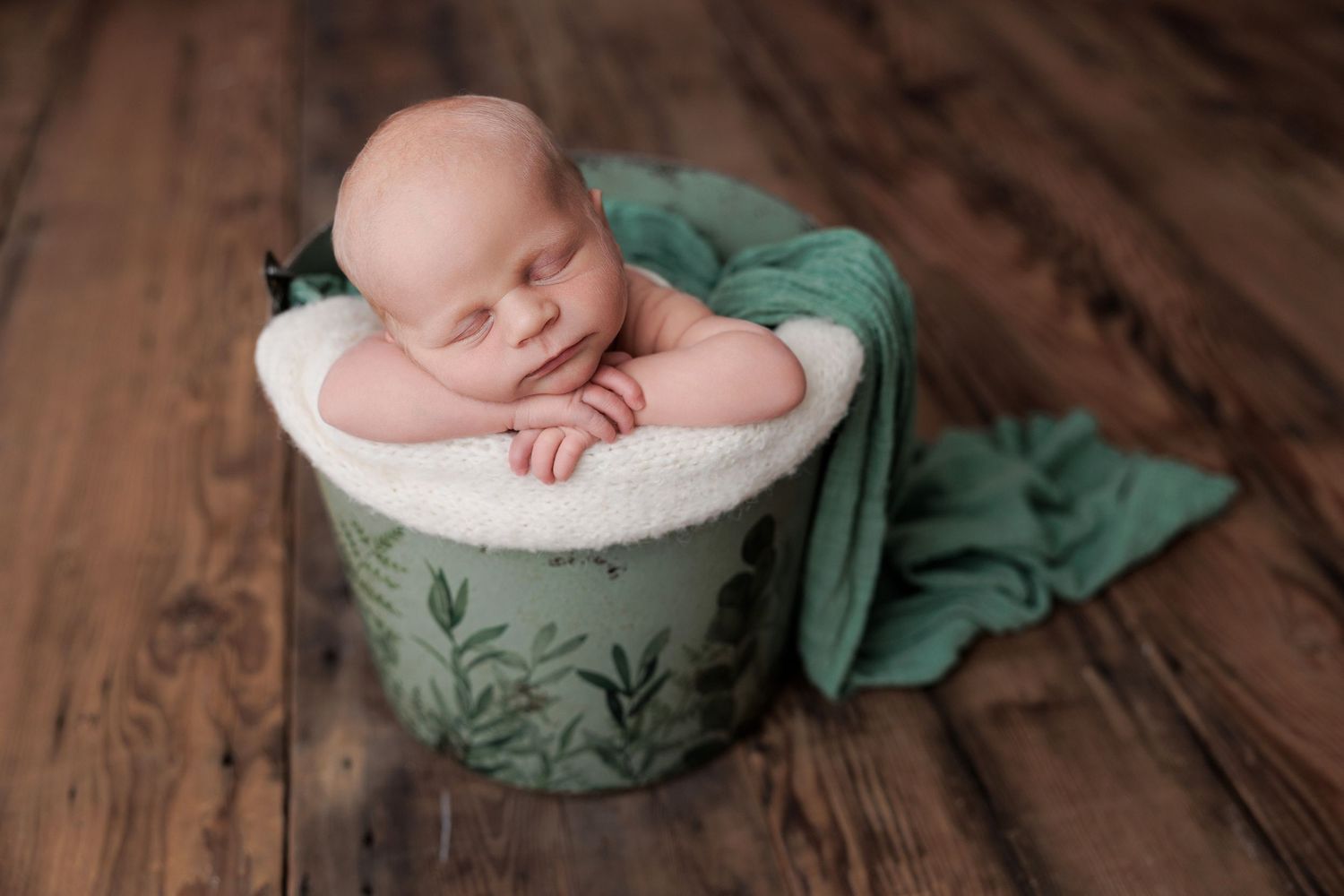 Sleeping newborn nestled in a decorative green and white container with sage fabric draping on wooden floor.