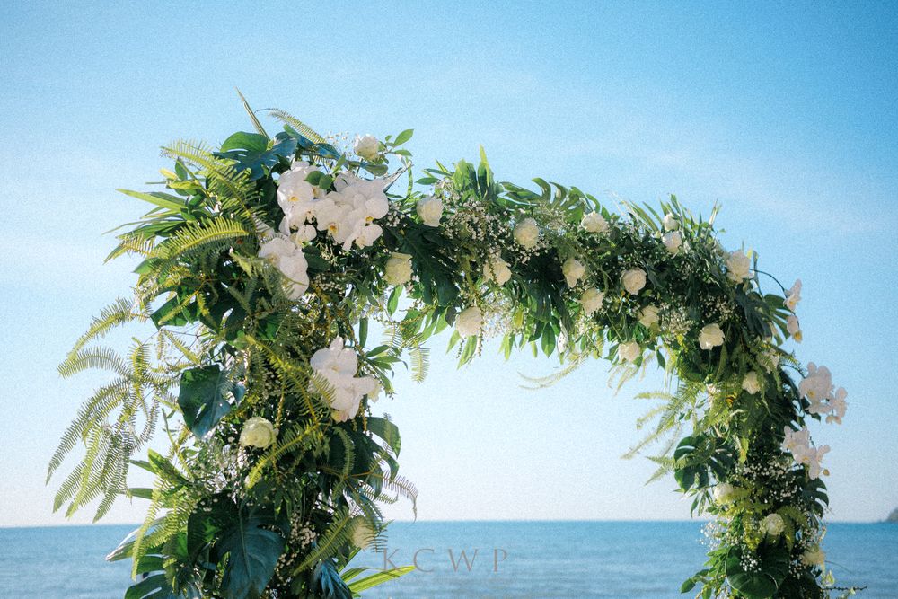 Wedding arch decorated with white flowers and lush greenery overlooks the ocean on a bright sunny day.