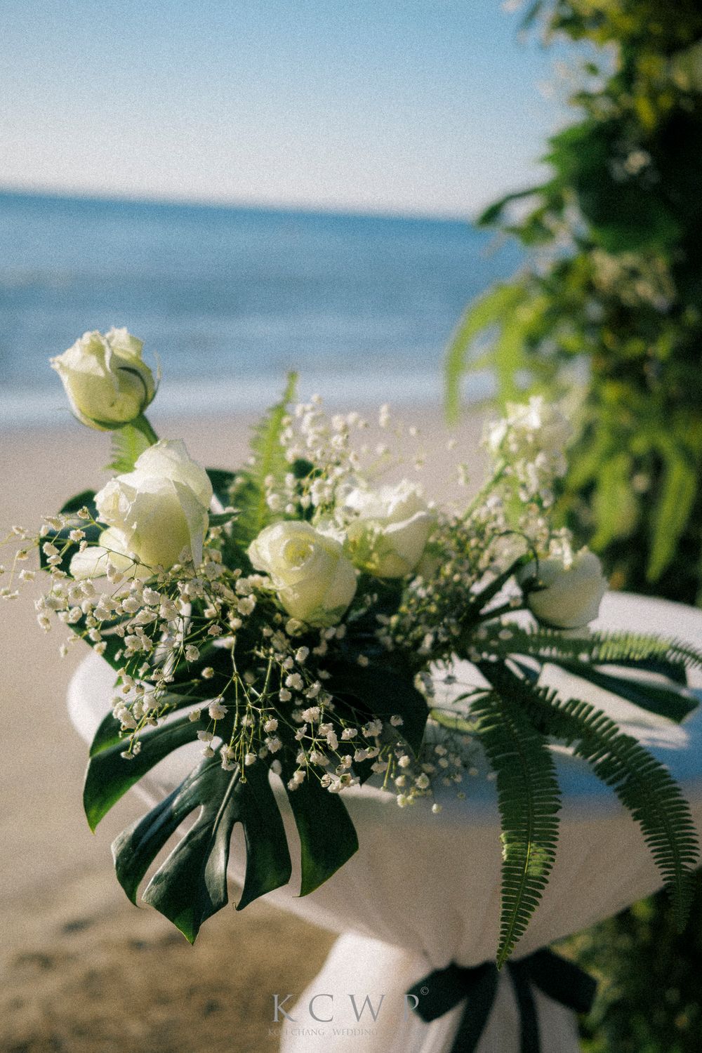 A tropical beach wedding floral arrangement with white roses, baby's breath, and monstera leaves against an ocean backdrop.