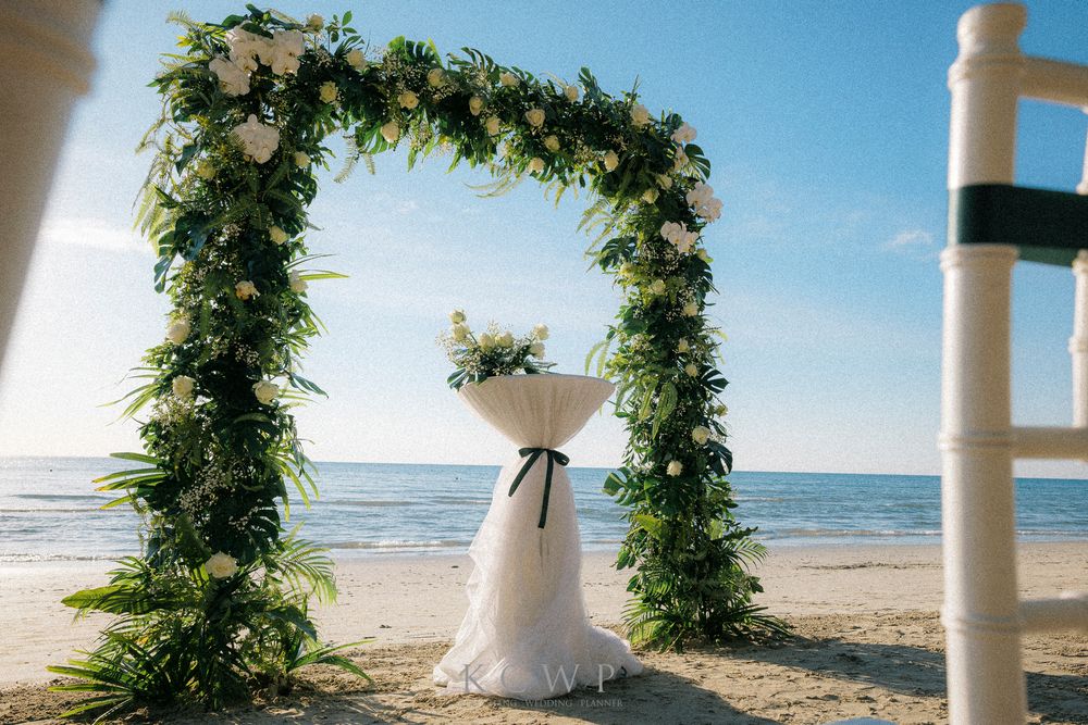 A floral wedding arch decorated with greenery stands on a sunny beach overlooking the ocean.