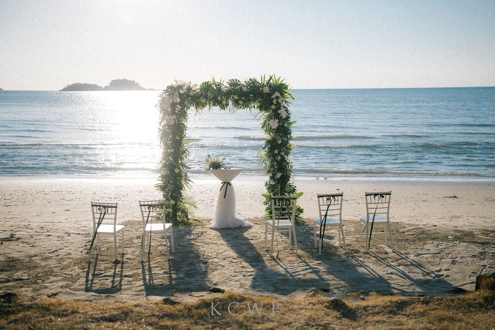 An elegant floral wedding arch with wooden chairs stands on a pristine beach overlooking the ocean on a sunny day.