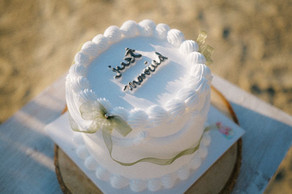 A small white frosted cake with light blue accents and delicate piped decorations sits on a pale surface.