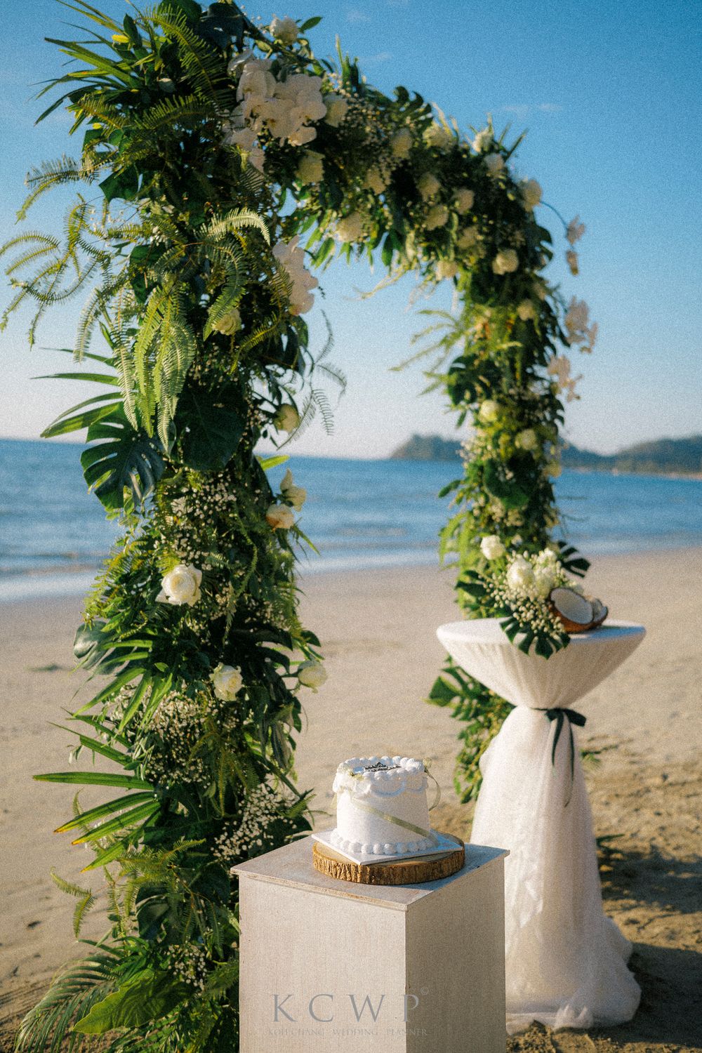 A lush floral wedding arch with white flowers and tropical greenery stands on a scenic beach with ocean views.