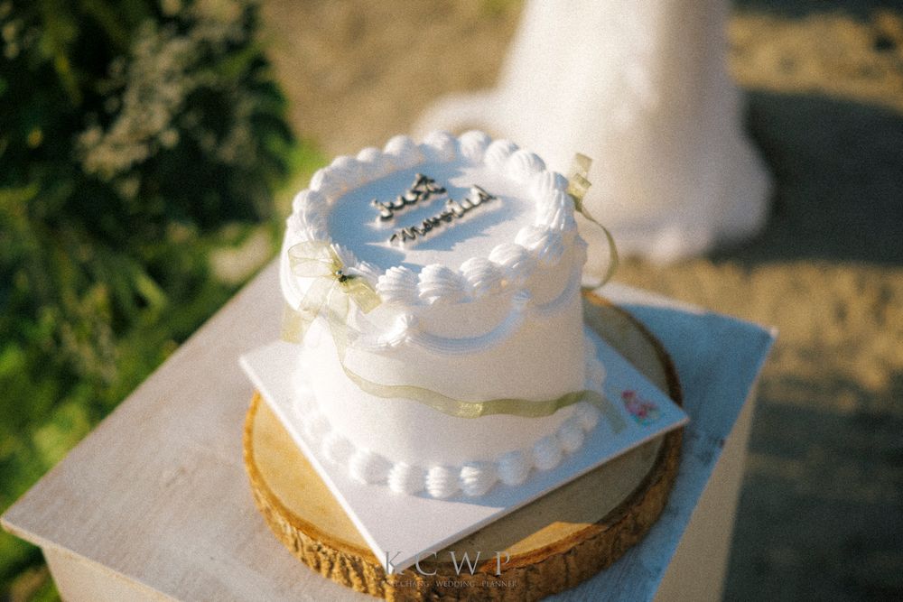 A detailed white two-tier wedding cake with delicate pearl piping decorations sits atop a gold base in outdoor lighting.