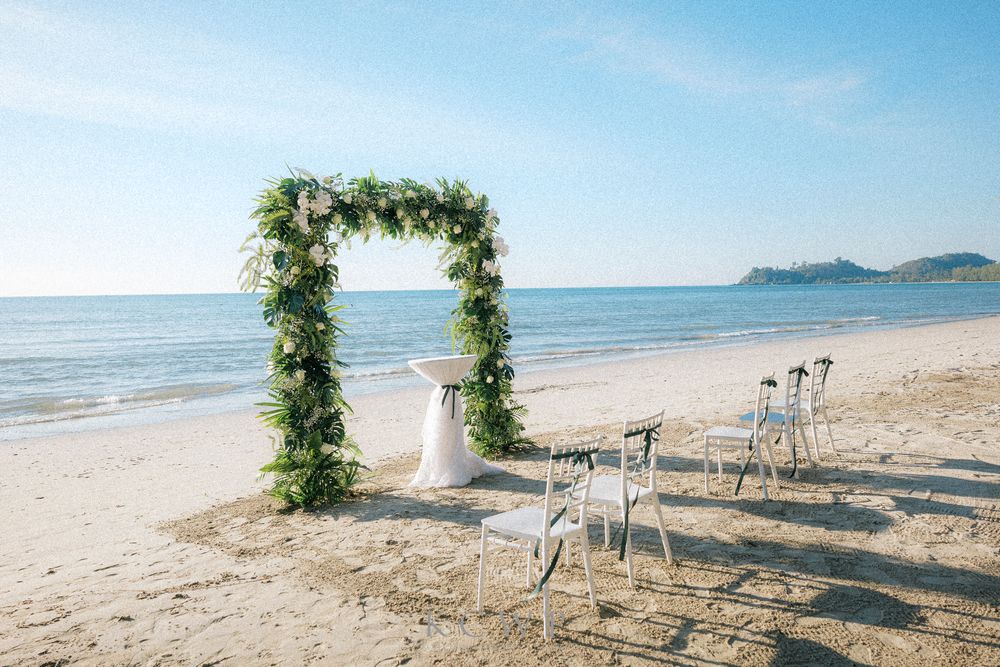 A floral wedding arch adorned with greenery stands on a pristine beach with white chairs and ocean views.