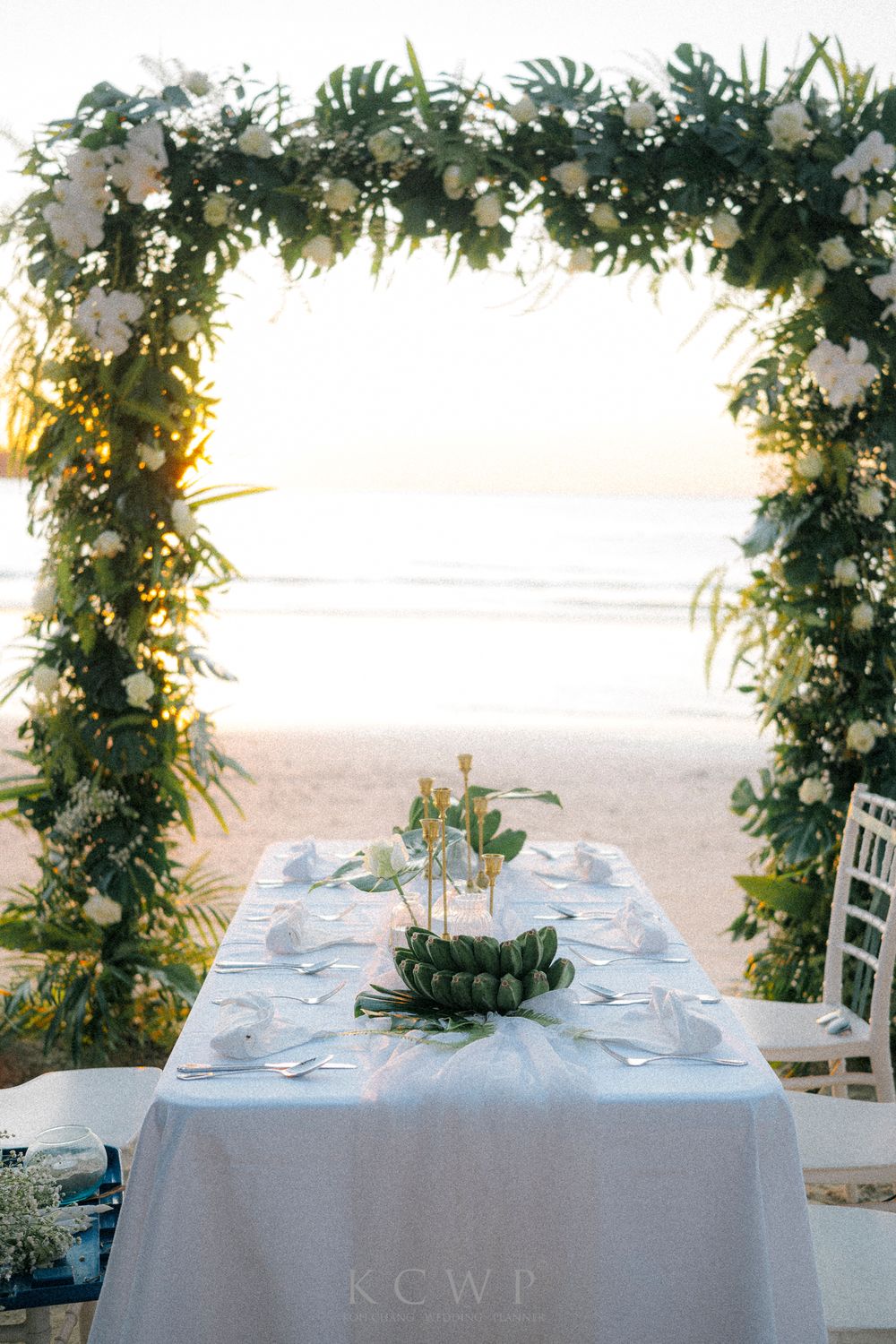 A romantic beach wedding table setting under a floral arch with white linens at sunset.