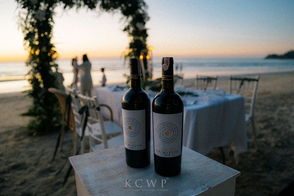 Wine bottles displayed on a white pedestal against a beach sunset with a floral arch in the background.